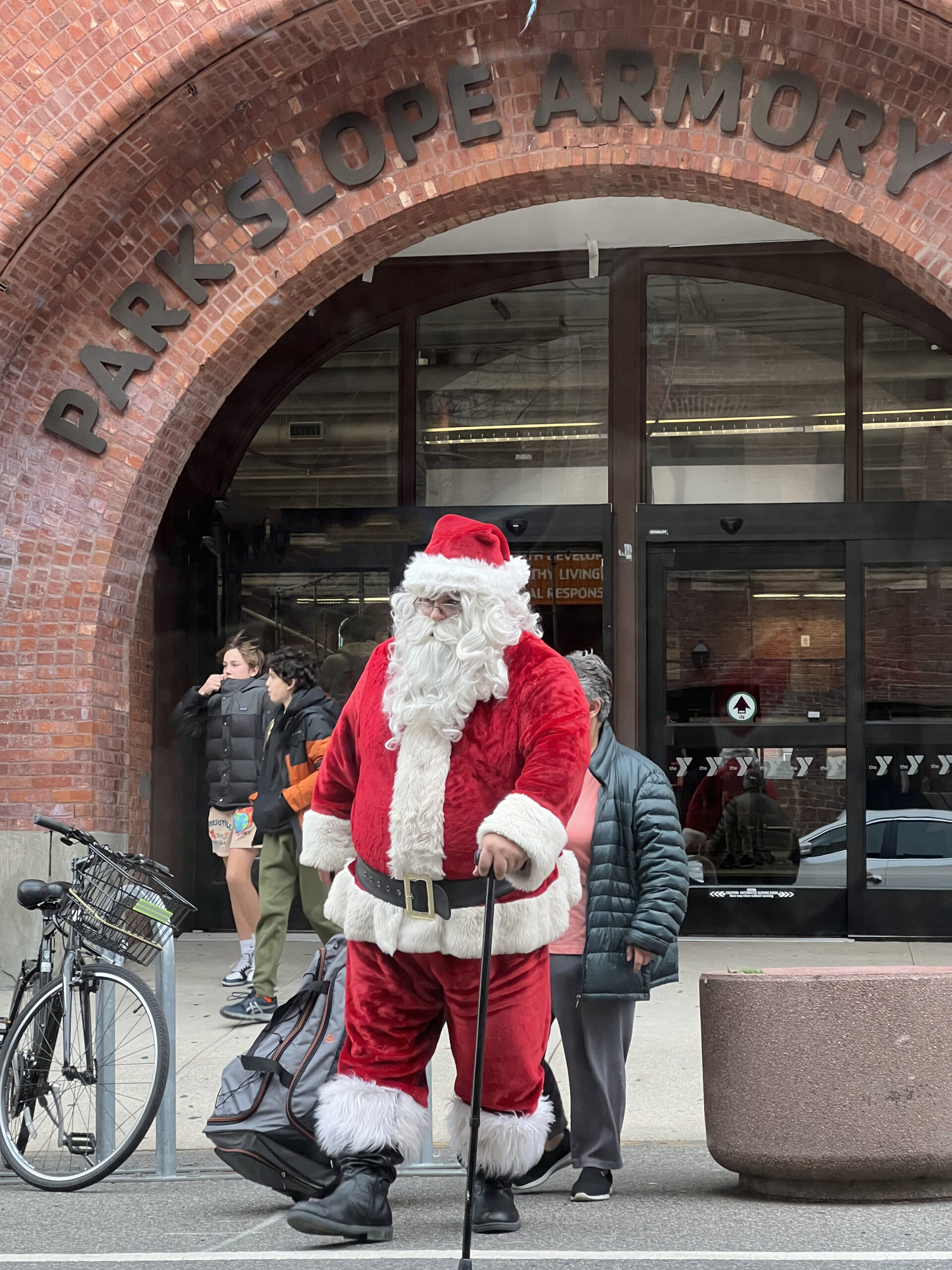 Photo of Santa Claus, in red coat with white trim, and large white beard, crossing a city street, walking with a cane.